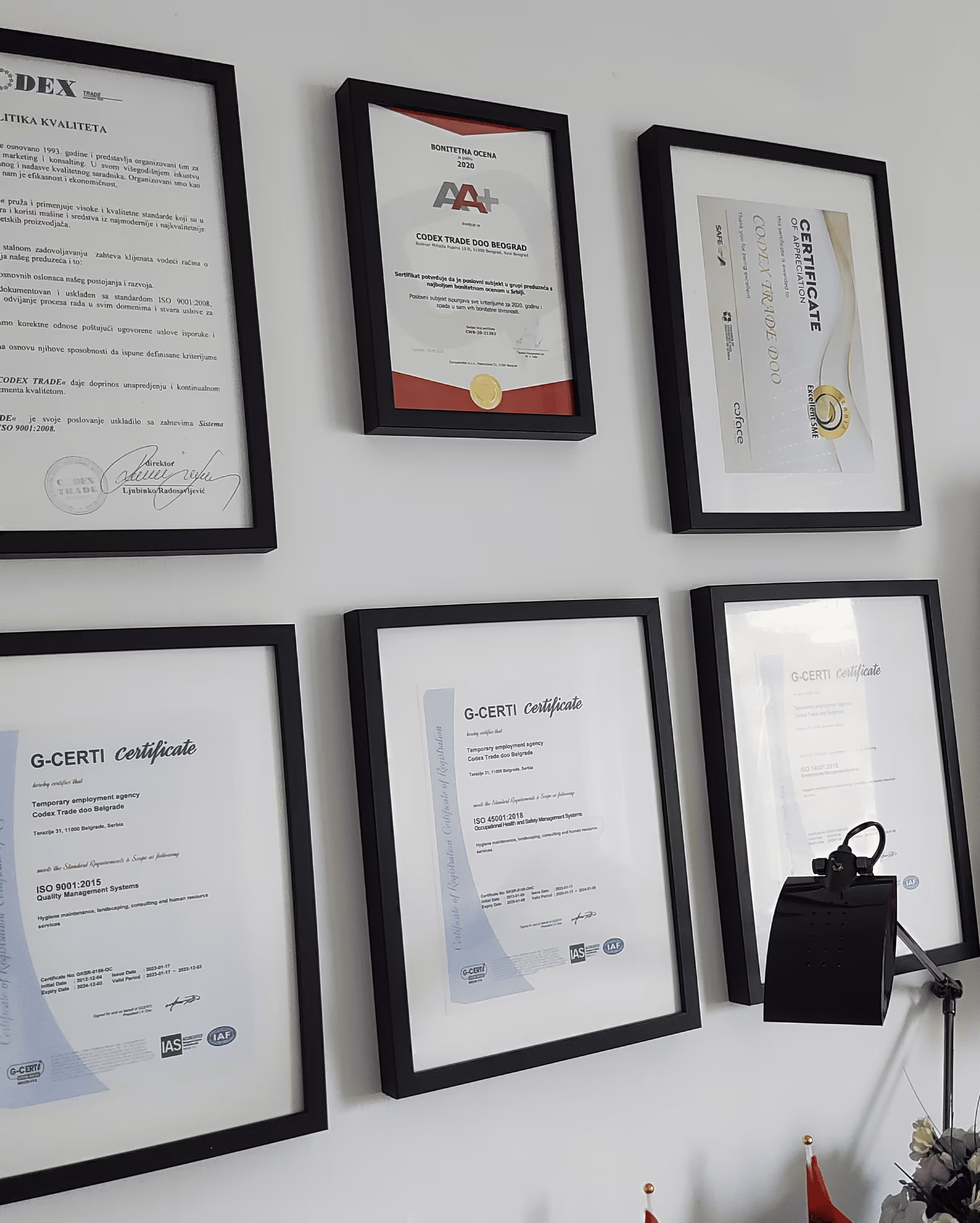 Six framed certificates and awards hanging on a white wall above a desk lamp.
